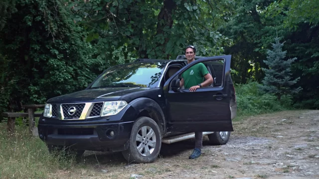 A man standing next to a black Nissan jeep in a forest setting.